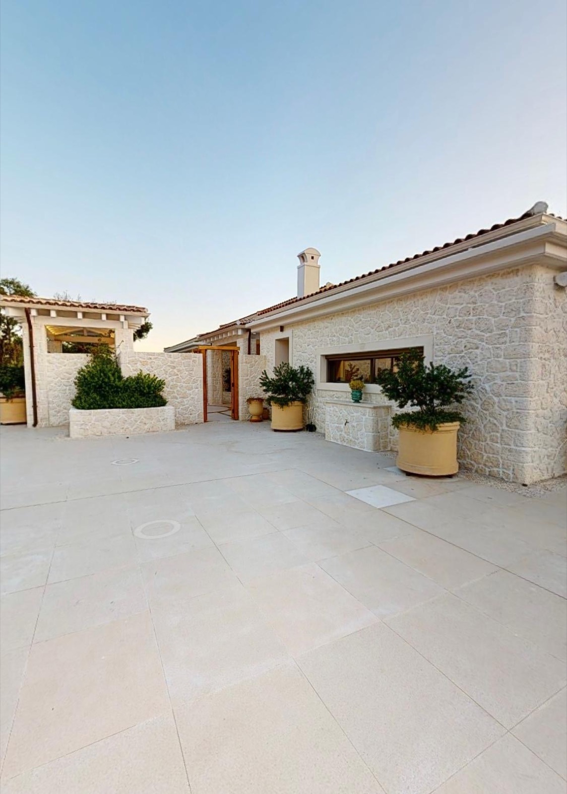 White polygonal stone cladding and a stone chimney in warm evening light, above a sand-toned paved courtyard with terracotta planters.