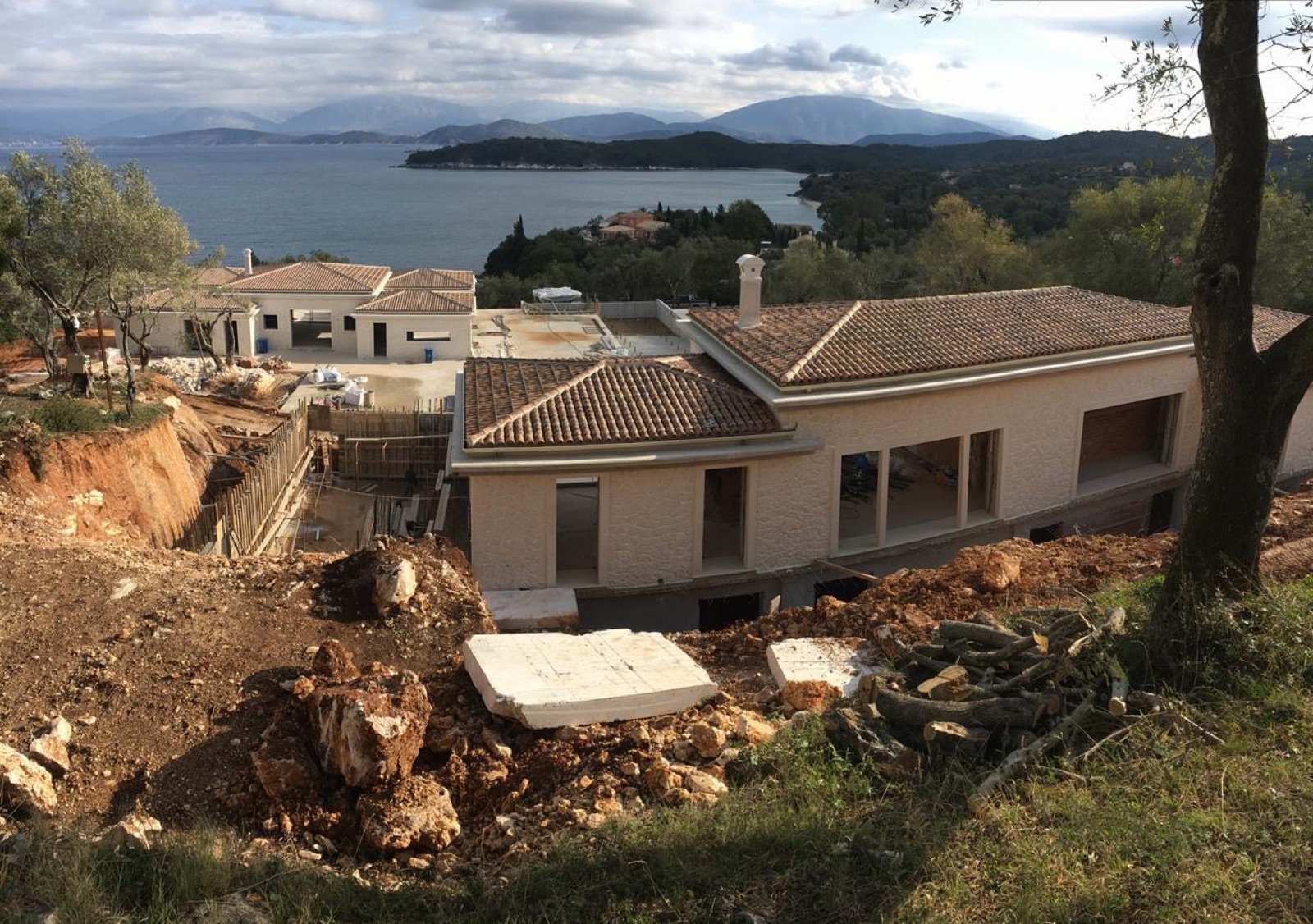 The roofed villa shell above raw earthworks, with a sweeping view over the bay, headlands and mountains under a dramatic sky.