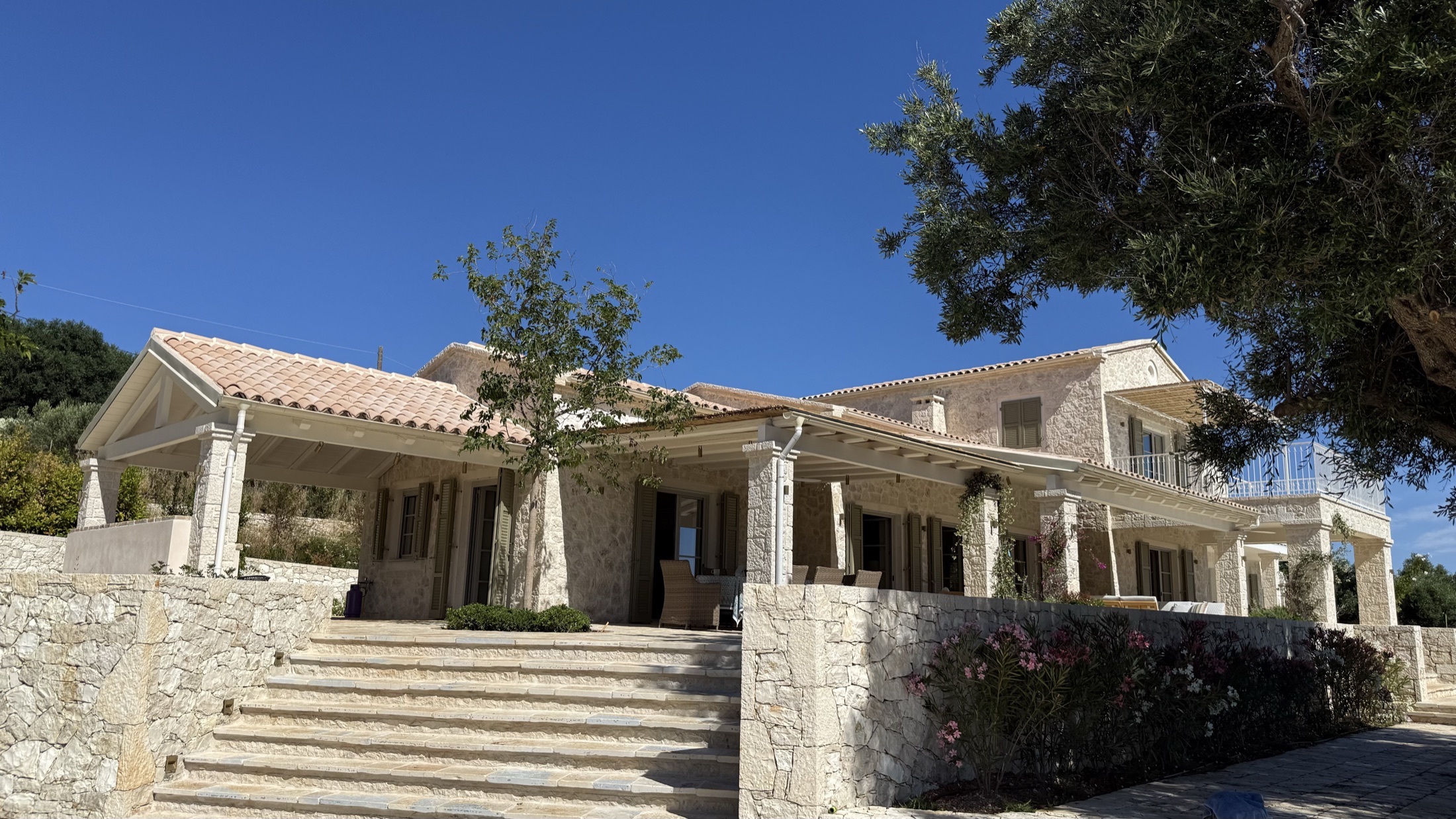 Broad stone steps rising to covered porches with stone columns, terracotta roofs, flowering beds and olive tree, cloudless blue sky