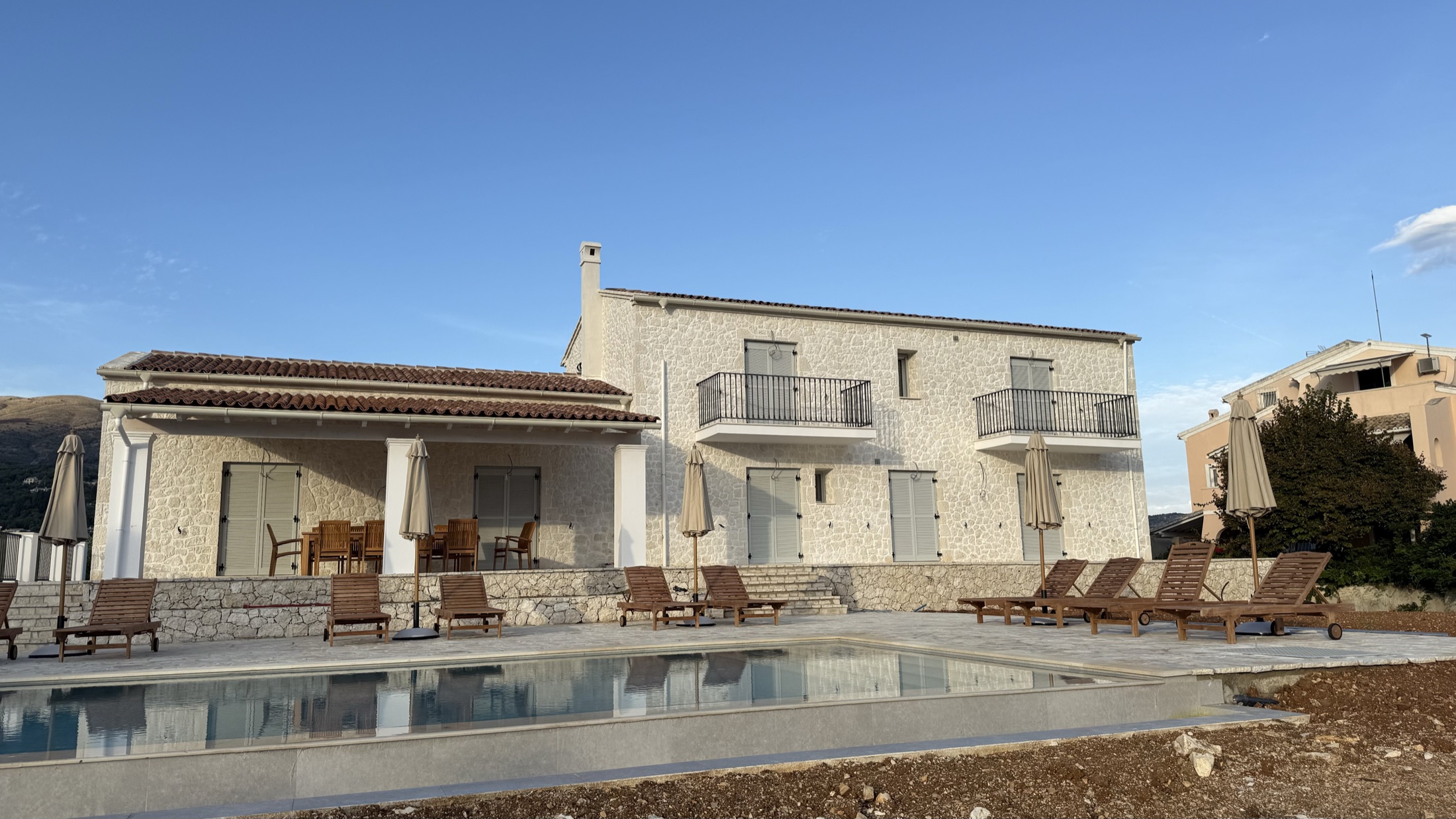 Full villa elevation in soft evening light with the infinity-edge pool reflecting the facade, lounge chairs and closed umbrellas along the stone-paved pool deck